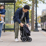 AI示意图:一名成年人在人行道上整理空的折叠婴儿车,另一名成年人抱着幼儿,背景是一辆没有标识的公交车。